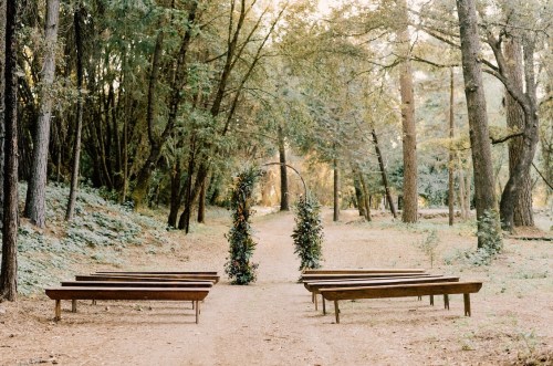 A serene outdoor wedding setup with wooden benches and a floral archway surrounded by trees.