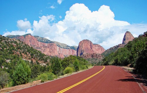 A winding road leads through a scenic landscape of red rock formations and lush greenery under a blue sky with clouds.