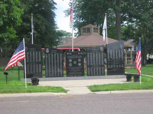 A memorial with black stone panels listing names, flanked by American flags, set in a park with a gazebo in the background.