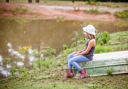 A young girl in a hat sits on a boat by a calm pond, surrounded by greenery and wildflowers.
