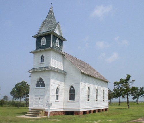 A small white church with a green roof and steeple, surrounded by grass and trees under a clear blue sky.