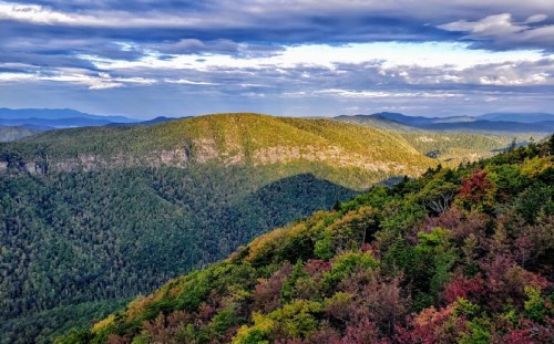 A panoramic view of lush green mountains under a cloudy sky, showcasing vibrant autumn foliage.