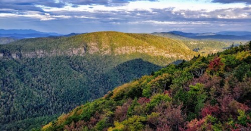 A panoramic view of lush green mountains with autumn foliage under a cloudy sky.
