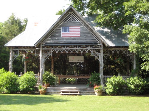 A rustic wooden pavilion with an American flag, surrounded by greenery and potted plants.