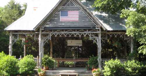 A rustic wooden pavilion with an American flag, surrounded by greenery and potted plants.