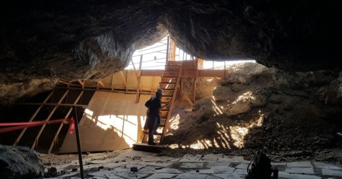 A person stands on a wooden ladder inside a cave, with light streaming through an opening and construction materials visible.