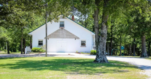 A white house with a triangular roof surrounded by trees and a grassy area, set in a peaceful outdoor environment.