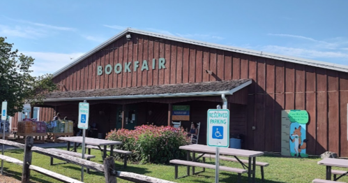 A rustic building labeled "BOOKFAIR" with outdoor seating and accessible parking signs in a sunny setting.