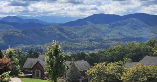 A scenic view of mountains and lush greenery, with houses in the foreground under a partly cloudy sky.