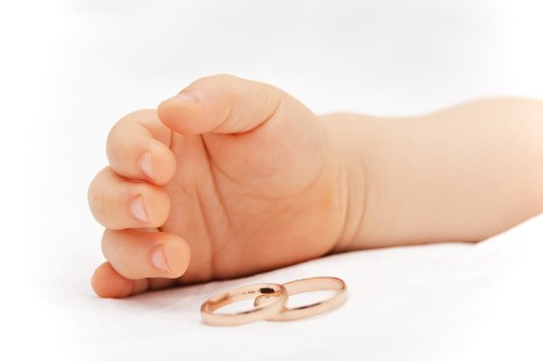 A close-up of a baby's hand reaching out next to two gold wedding rings on a white surface.