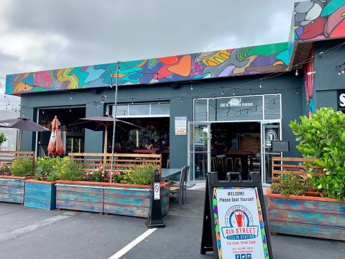 Colorful mural on a restaurant exterior with outdoor seating and a welcome sign in front.