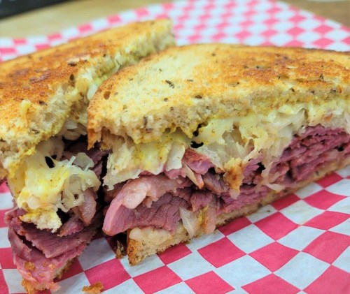 Close-up of a toasted sandwich filled with layers of corned beef and sauerkraut, served on a red and white checkered paper.