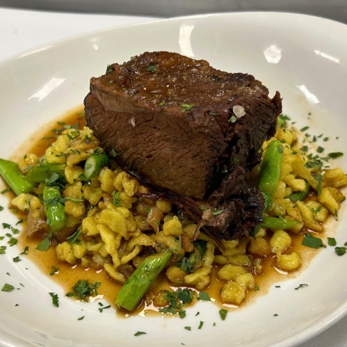 A plate of tender beef on a bed of spätzle, garnished with green asparagus and herbs in a savory sauce.