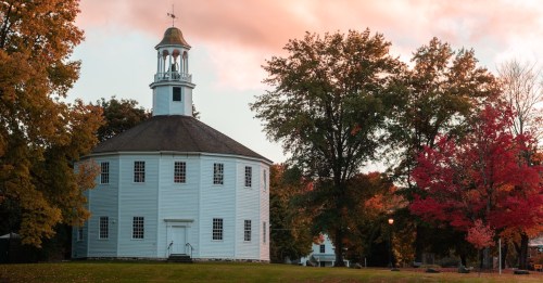 Historic octagonal Old Round Church in Richmond, VT, surrounded by vibrant fall foliage at sunset.