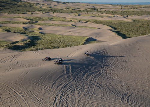 Three off-road vehicles navigate sandy dunes under a clear sky, leaving tire tracks in the soft sand.