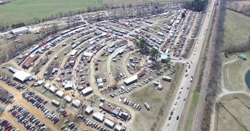 Aerial view of a large outdoor market with winding rows of stalls and parked cars, surrounded by green fields.