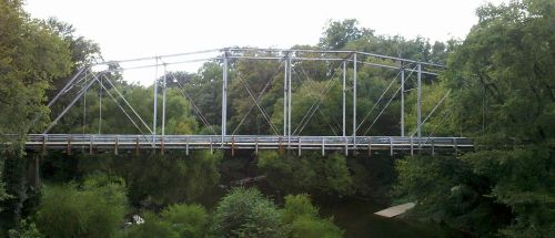 A metal truss bridge spans a lush, green riverbank surrounded by trees.