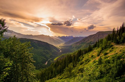 A breathtaking valley view at sunset, with mountains, lush greenery, and a winding river under a colorful sky.