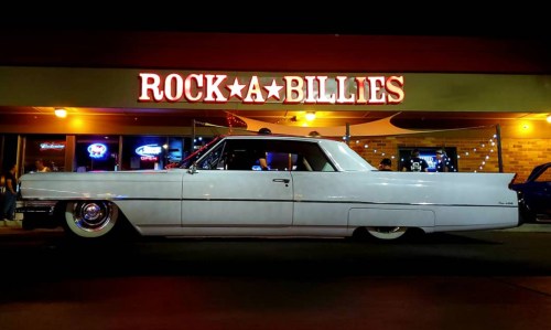 A classic white car parked in front of a neon-lit diner named "Rock-A-Billies" at night.