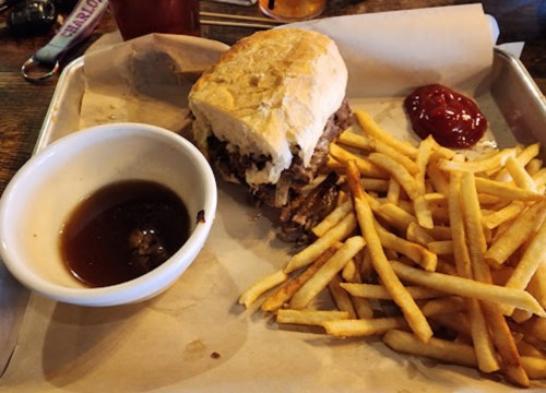 A sandwich filled with meat on a bun, served with a side of fries and a small bowl of dipping sauce.