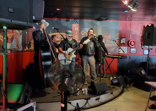 A band performs on stage in a lively venue, featuring a guitarist, singer, and keyboardist with colorful lighting.