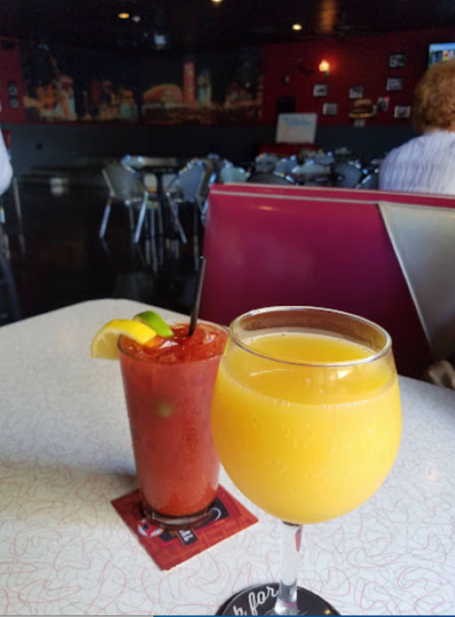 Two cocktails on a table: a Bloody Mary with a lime wedge and a mimosa in a glass, set in a casual dining space.