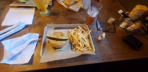 A tray with a burger topped with a pickle and a side of fries, surrounded by drinks and condiments on a wooden table.