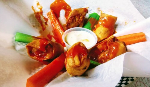 A plate of crispy spring rolls drizzled with sauce, surrounded by celery and carrot sticks, with a dipping sauce in the center.