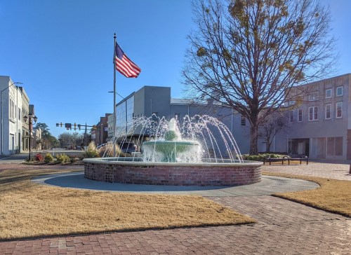 A fountain with water jets in a plaza, with an American flag flying nearby and buildings in the background.
