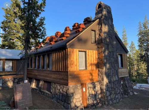 A rustic house with a stone exterior and wooden accents, surrounded by trees and a clear blue sky.