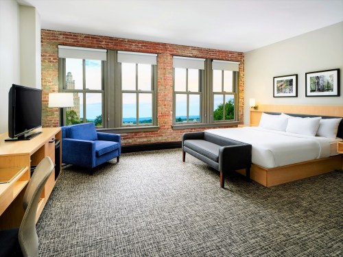 Modern hotel room with a large bed, blue armchair, desk, and windows showcasing a scenic view.