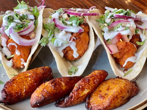 Three tacos filled with fried fish, topped with onions, cilantro, and sauce, served with golden fried plantains.