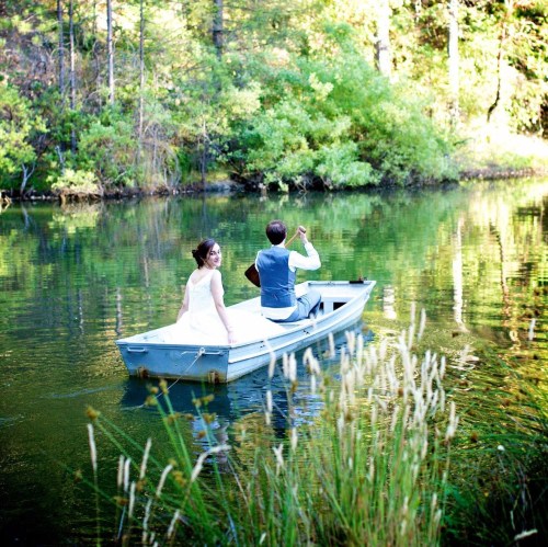 A bride and groom in a small boat on a serene lake, surrounded by lush greenery and reflecting water.