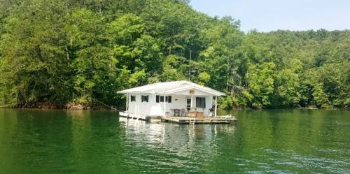 A small white house on a floating dock surrounded by lush green trees and calm water.
