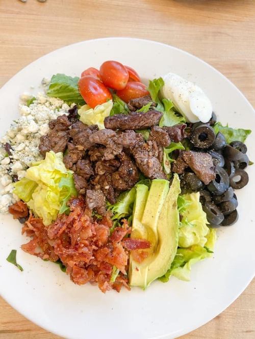 A colorful salad with lettuce, grilled steak, tomatoes, olives, avocado, bacon, and blue cheese on a white plate.