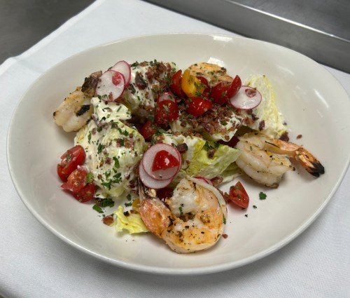 A plate of grilled shrimp on lettuce, topped with cherry tomatoes, radishes, and a creamy dressing, garnished with herbs.