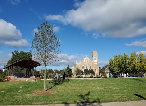 A park scene featuring a grassy area, a tree, a stage, and a historic building under a blue sky with clouds.