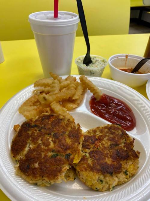 A plate with two golden crab cakes, crinkle fries, ketchup, coleslaw, and a drink on a bright yellow table.