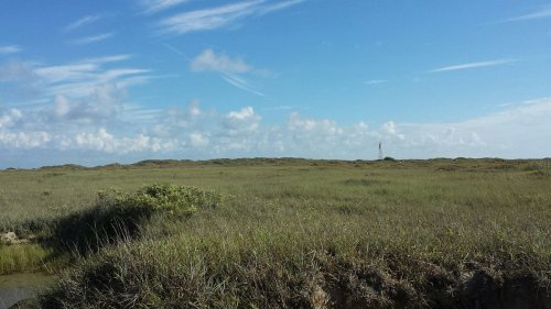 A wide view of a grassy landscape under a blue sky with scattered clouds and a distant structure on the horizon.