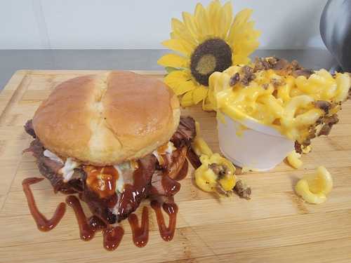 A BBQ sandwich with sauce on a wooden board, accompanied by a cup of mac and cheese topped with crumbled bacon and a sunflower.