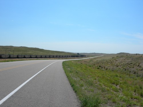 A long, empty road stretches through grassy plains under a clear blue sky, with hills in the distance.