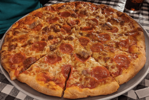 A large pepperoni and sausage pizza on a round tray, sliced into eight pieces, on a checkered tablecloth.