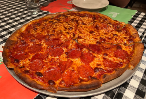A large pepperoni pizza with melted cheese, sliced into wedges, on a checkered tablecloth.