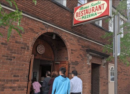 A group of people waiting in line outside Mama Santa's Restaurant and Pizzeria, featuring a brick facade and a colorful sign.