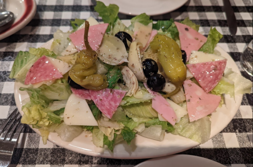 A colorful salad with lettuce, olives, pepperoncini, and various sliced meats and cheeses on a checkered tablecloth.