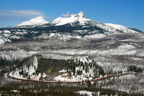 Snow-covered mountains rise above a forested landscape, with a winding road visible in the foreground.
