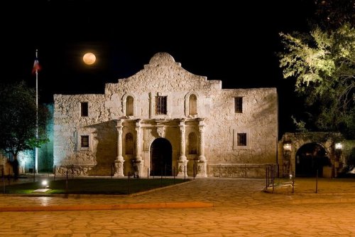 The Alamo illuminated at night, with a full moon in the sky and trees nearby.