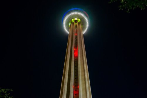 A tall tower illuminated with colorful lights at night, featuring a circular observation deck at the top.