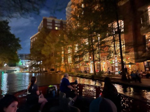 A nighttime scene of people on a boat by a lit-up canal, with trees and buildings reflecting in the water.