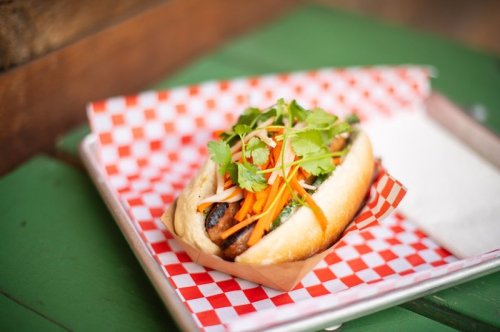 A gourmet hot dog topped with carrots, cilantro, and other fresh ingredients, served on a checkered paper.